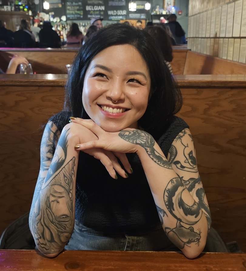 A smiling woman with black hair and tattoos on both arms sitting at a restaurant table. She has her hands under her chin and is looking to the left. The background features a busy restaurant or bar with people and a menu on the wall.