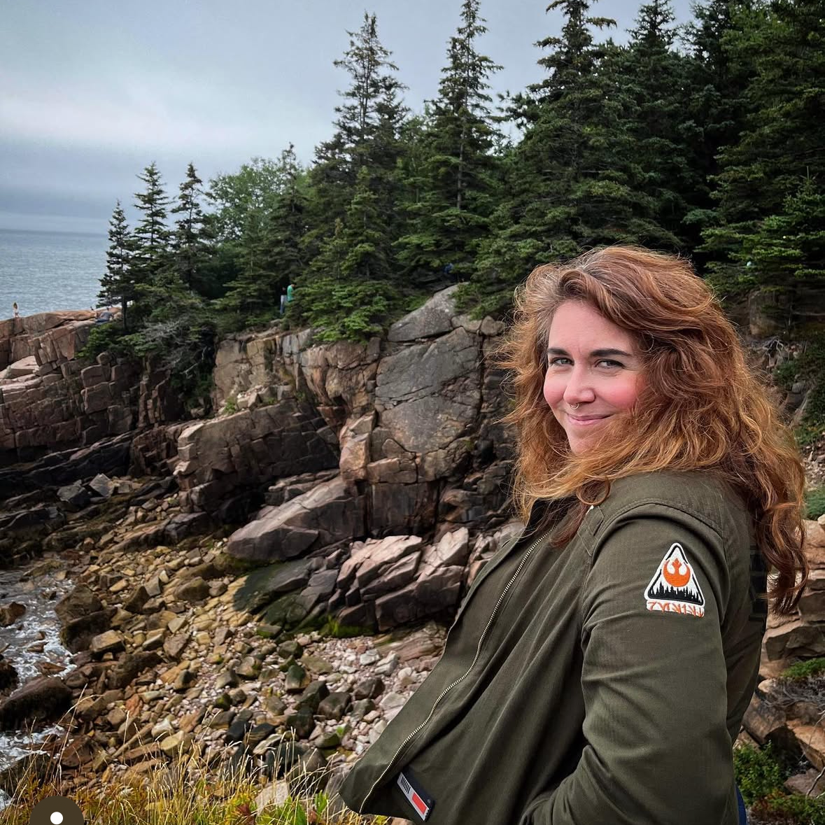A woman with wavy red hair and a piercing smile stands outdoors near rocky terrain and a dense forest of pine trees, with a cloudy sky overhead.