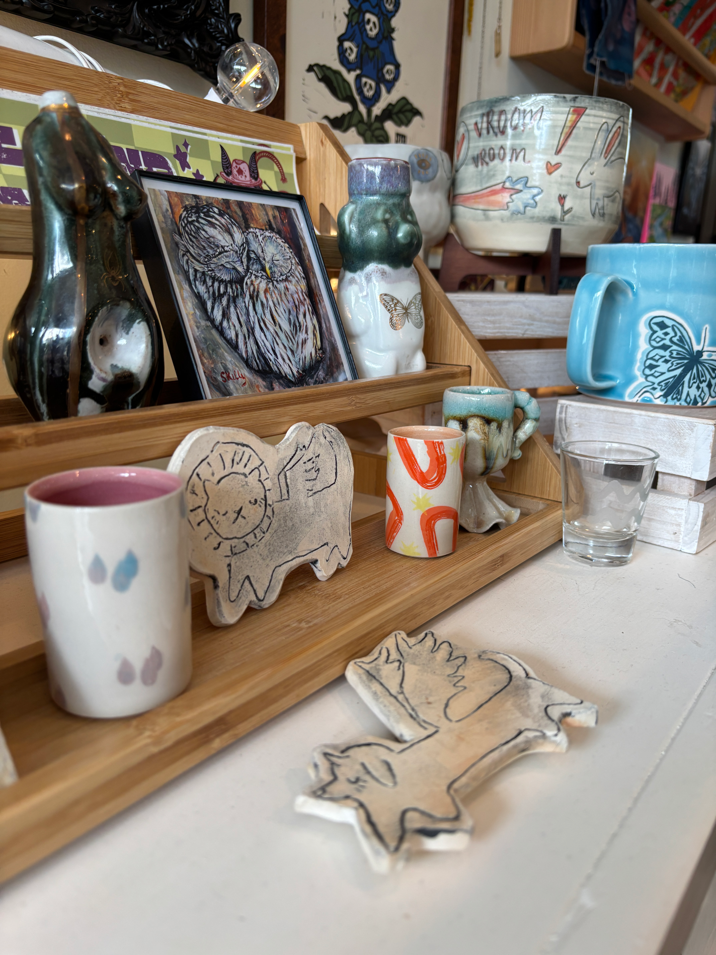 Display of various handmade ceramic items, including cups, small sculptures, and decorative objects, arranged on a wooden tray and shelf in a store.