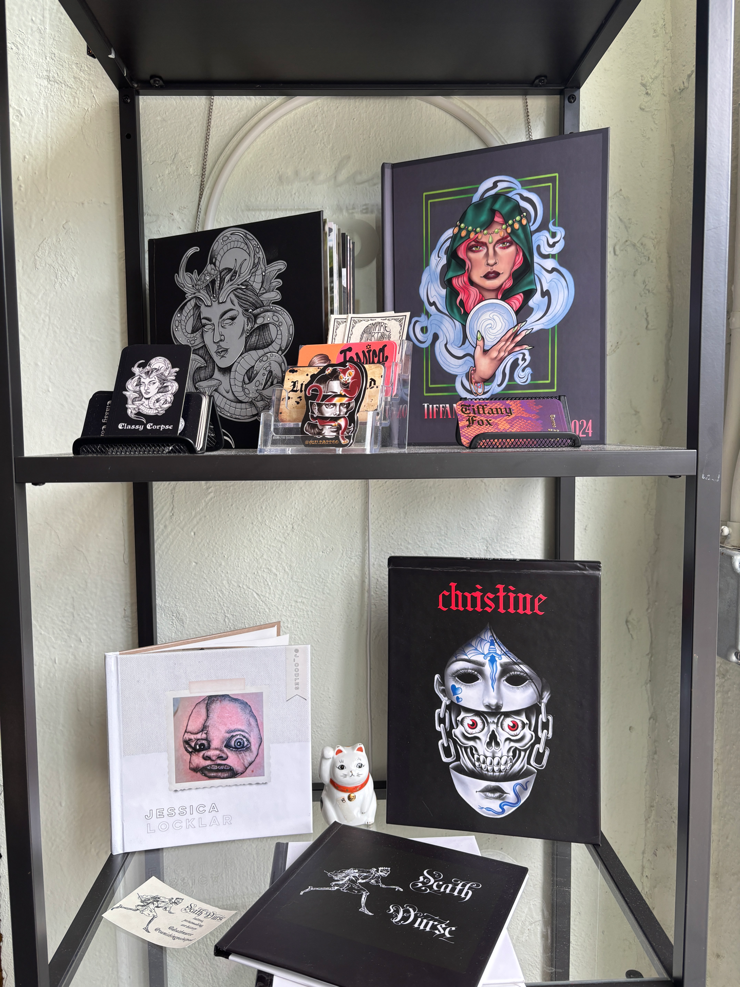Black metal shelving unit displaying various artworks and prints, including a framed black and white illustration of a woman with snakes around her head, a colorful portrait of a woman with a hood, and art books. A white sheet with a child's drawing, a white lucky cat figurine, and a black book titled 'Earth & Noise' are also on the shelves.
