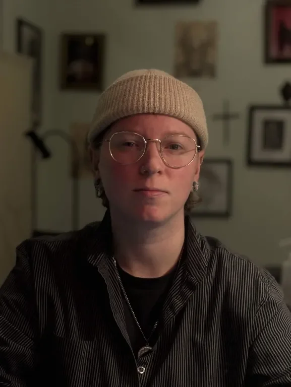 A young person with short hair, wearing glasses, a beige beanie, and a dark striped shirt, sitting in a room with framed pictures on the wall.