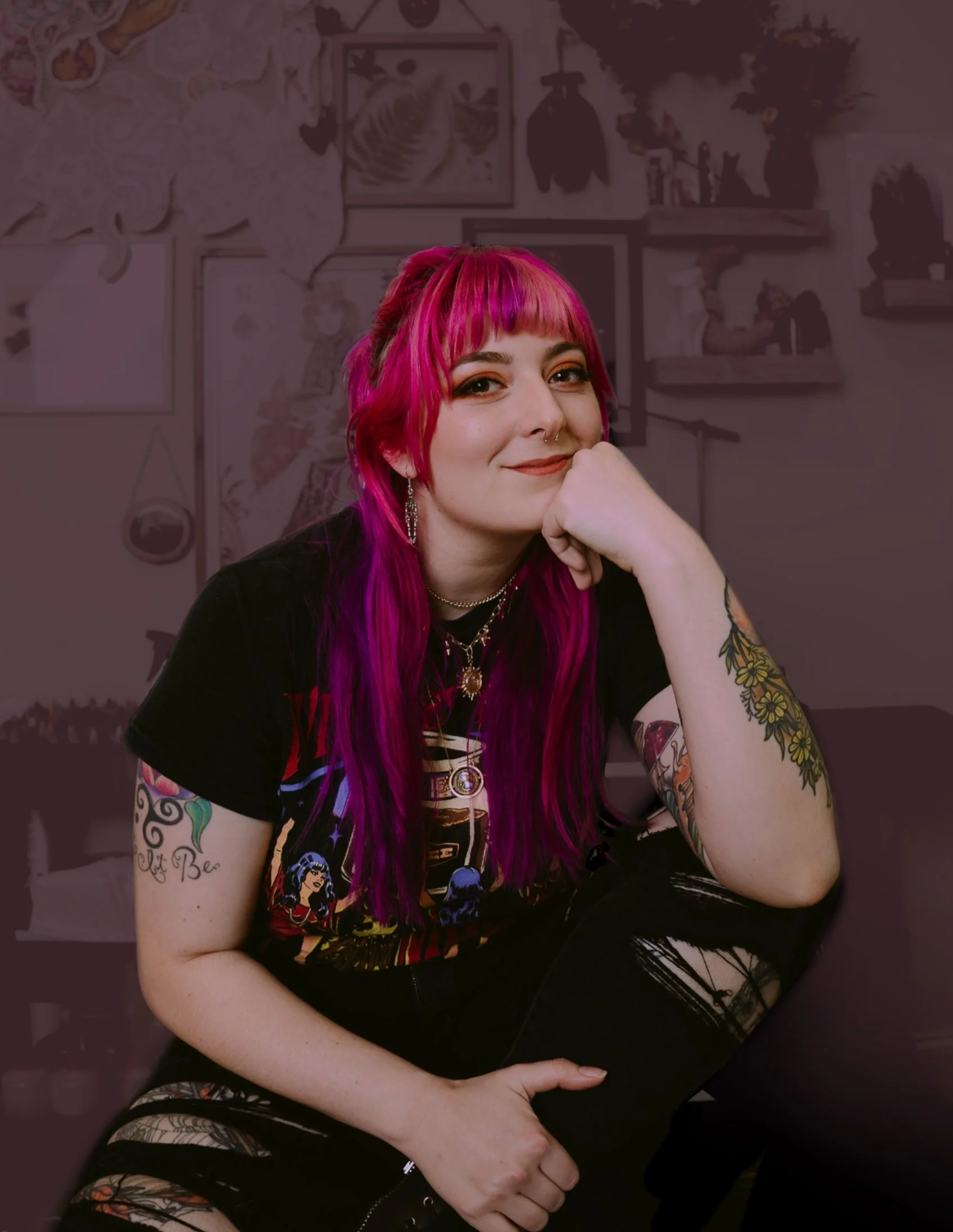 A woman with vibrant pink hair, multiple tattoos, and colorful jewelry sitting in front of a wall decorated with framed art and plants.
