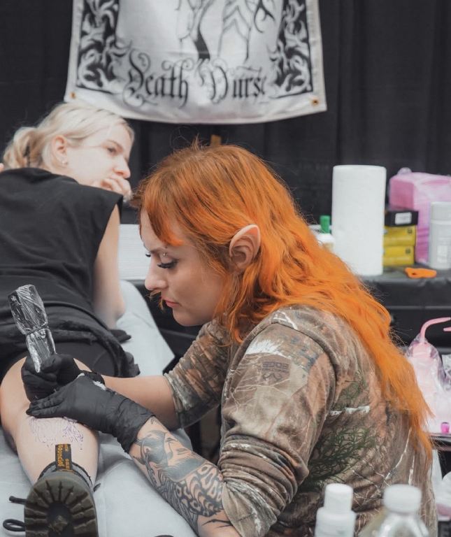 A woman with orange hair, tattoos, and a camouflage shirt getting a tattoo done on her arm by an artist with platinum hair and black gloves in a tattoo studio. There is a banner with a gothic design and the words 'Death Vurs' in the background.