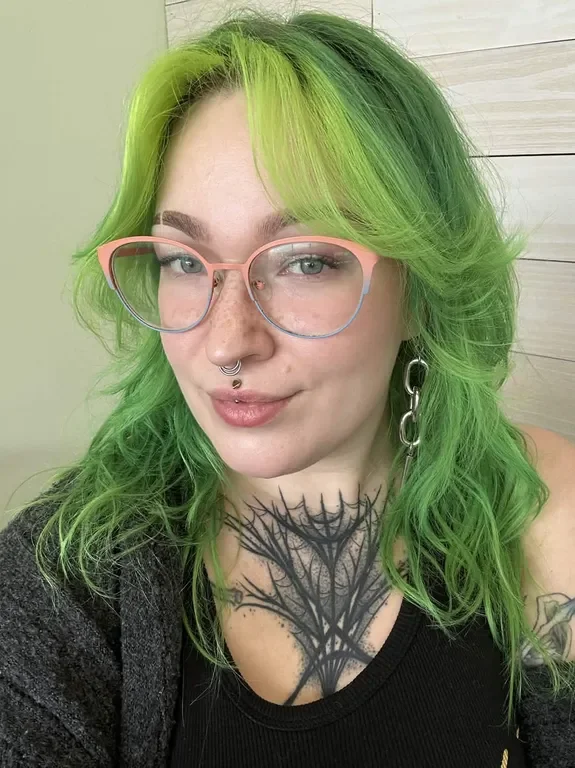 Woman with green hair wearing pink and blue glasses, large dangling earrings, a black tattoo on her chest resembling a spider web, and a septum piercing.