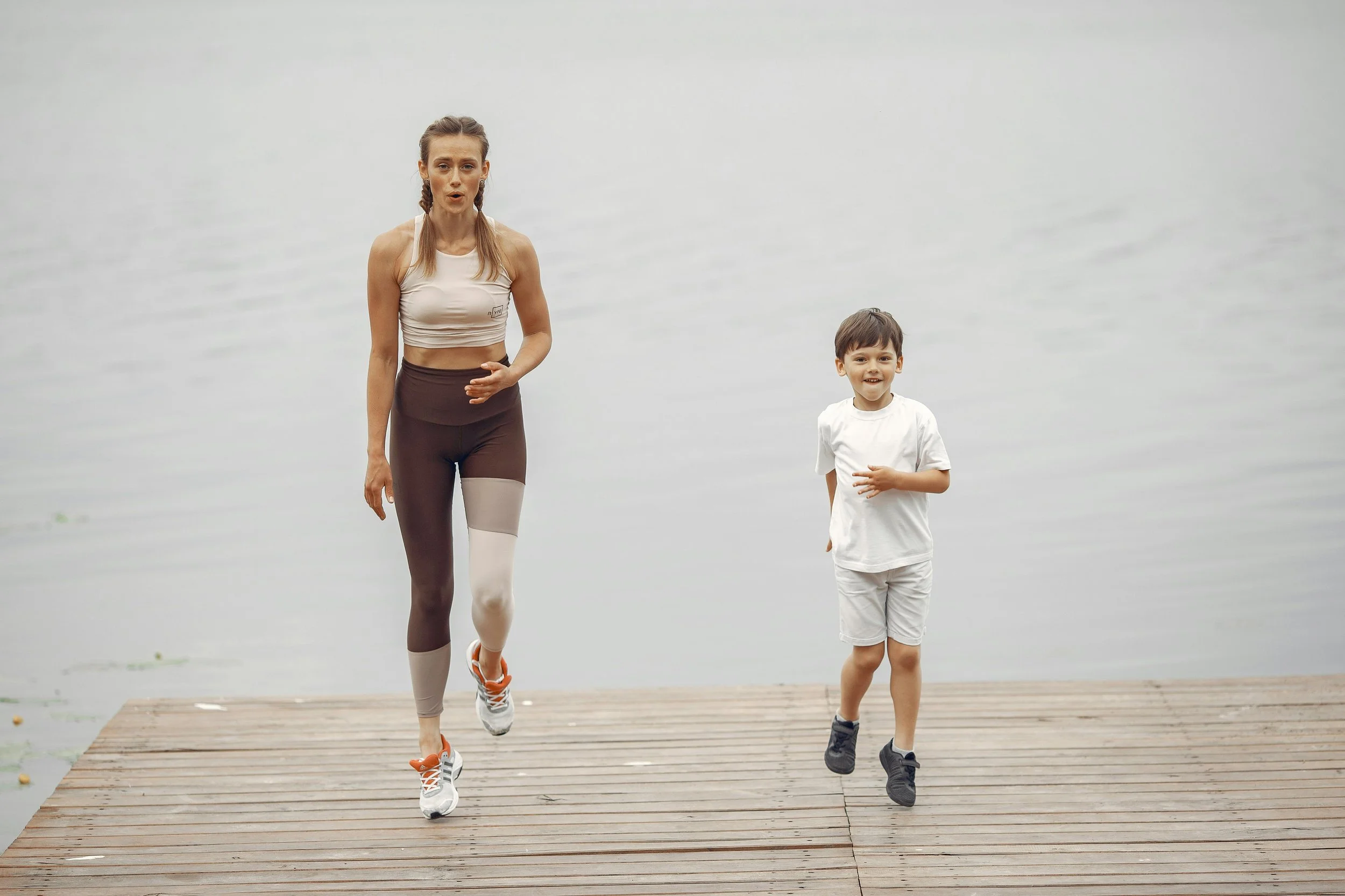 A woman and a young boy are jogging on a wooden dock by the water, both smiling and wearing athletic clothing.
