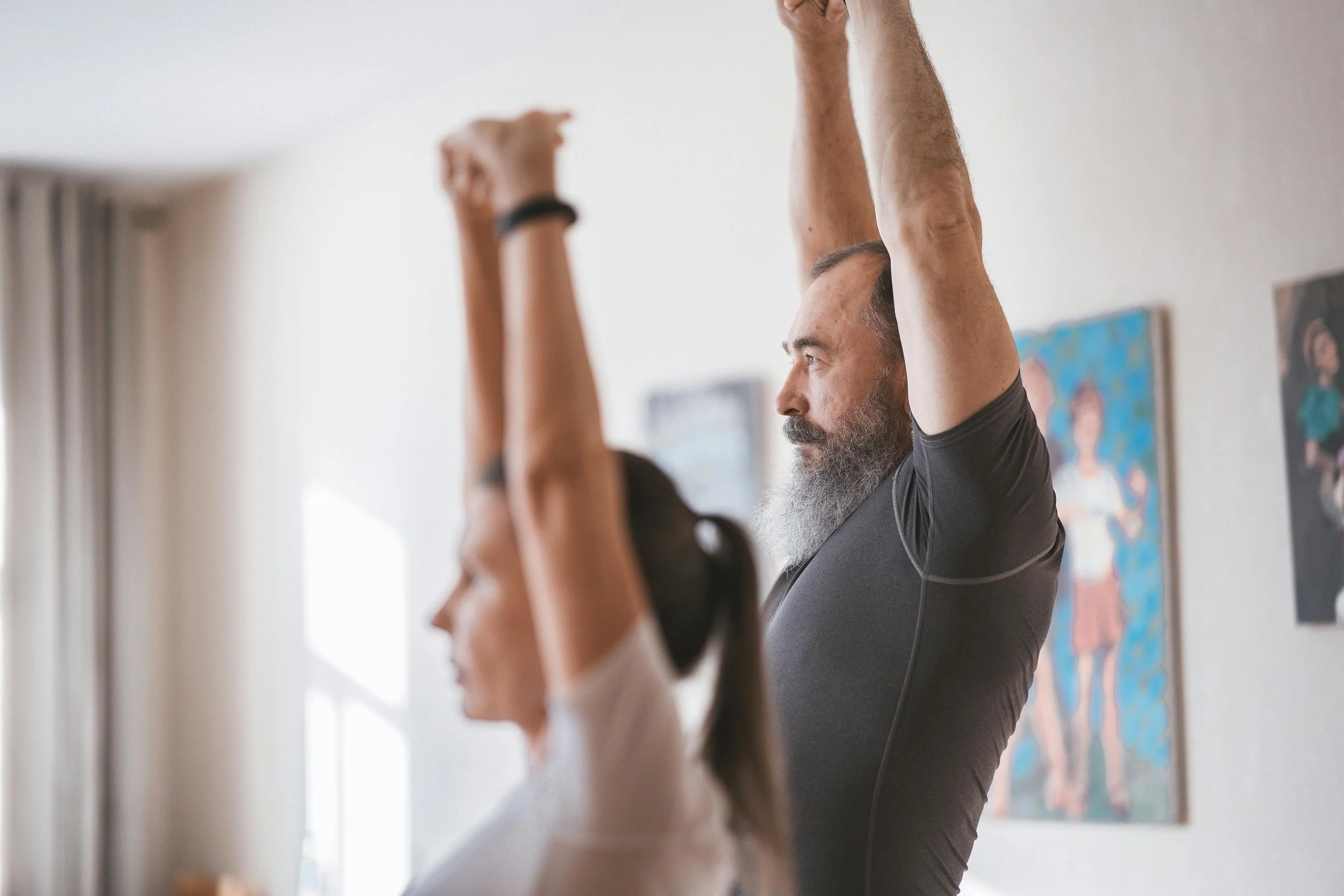 A man and woman in a room stretching with their arms raised overhead, facing sideways. The man has a beard and gray hair, wearing a black shirt. The woman has long dark hair and is wearing a white shirt. The room has light-colored walls decorated with framed artwork.