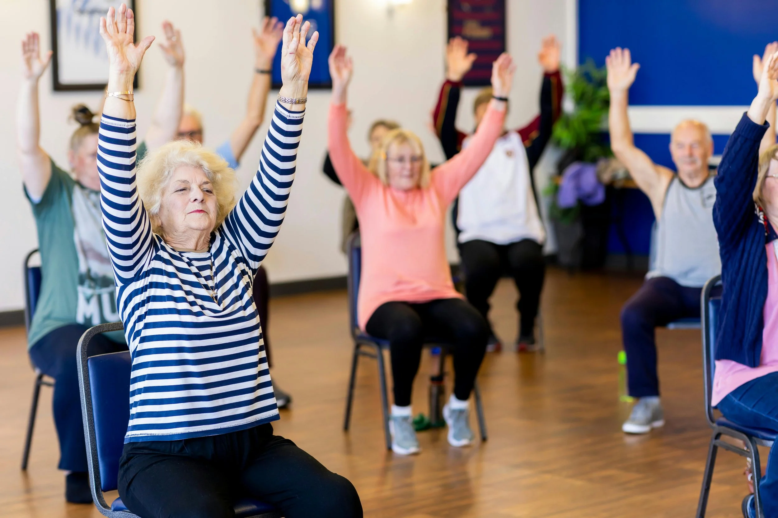 Group of older adults participating in a seated exercise class, raising their arms overhead.