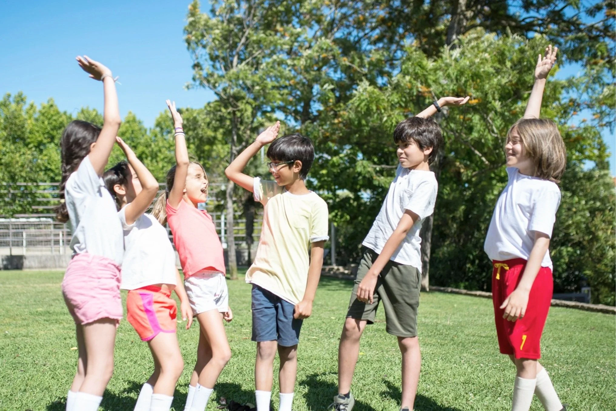 Group of children playing flag football on a grassy field during daytime, raising their hands happily.