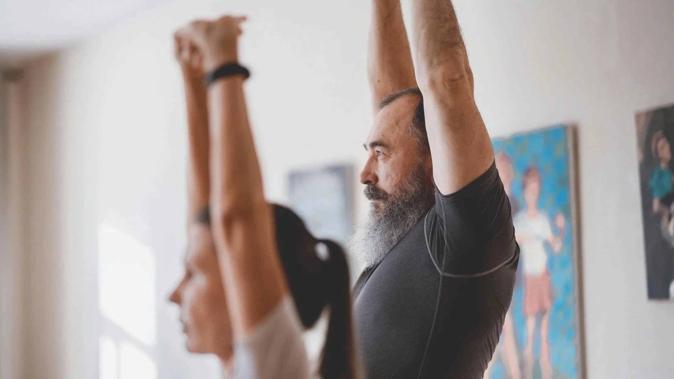 A man and woman in a room stretching with their arms raised overhead, facing sideways. The man has a beard and gray hair, wearing a black shirt. The woman has long dark hair and is wearing a white shirt. The room has light-colored walls decorated with framed artwork.