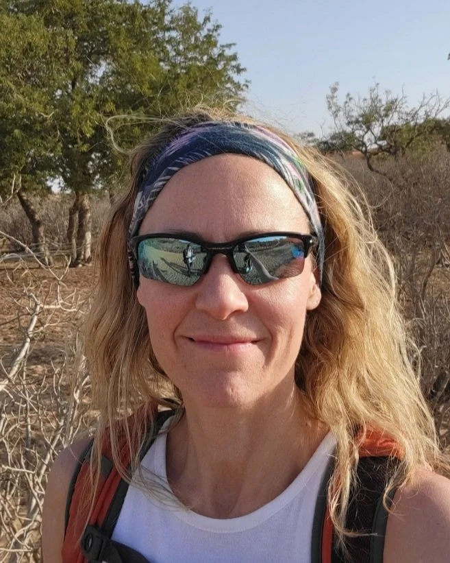 A woman taking a selfie outdoors in a dry, wooded area with leafless branches and green trees in the background. She is wearing sunglasses, a colorful headband, a white sleeveless top, and a backpack.