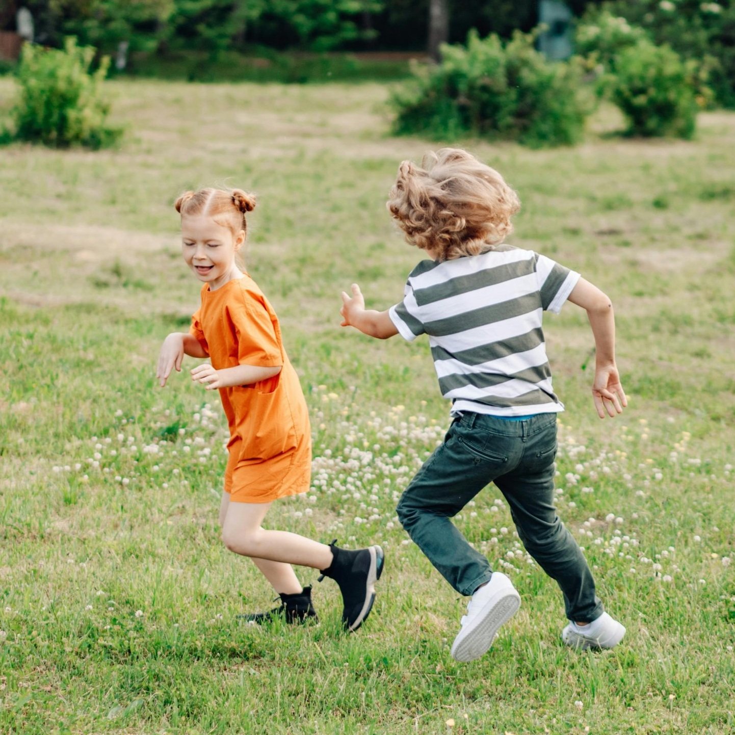 Two children, a girl in an orange dress and a boy in a striped shirt, are playing and running on a grassy field outdoors.