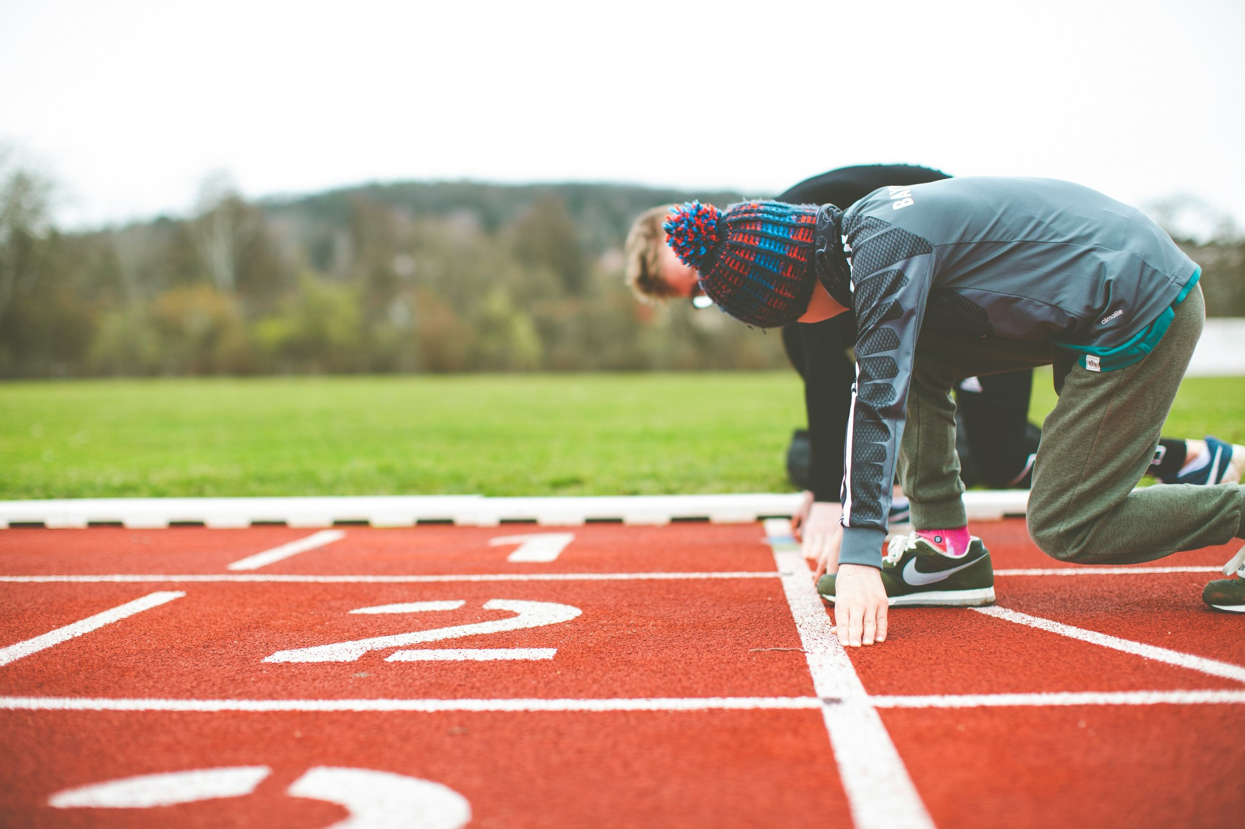 People in athletic clothing preparing for a race on an outdoor running track, positioned in starting blocks, with a background of grass and trees.