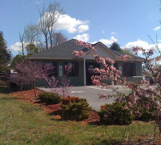 A small suburban house with a gray exterior and a dark roof, surrounded by blooming pink and white flowering trees, with a clear blue sky in the background.
