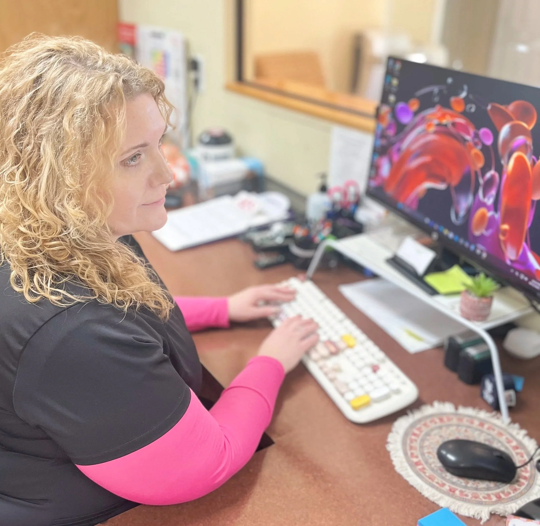A woman with curly blonde hair typing on a white keyboard at a desk, with computer monitor displaying colorful abstract art, various office supplies, and a small potted plant.