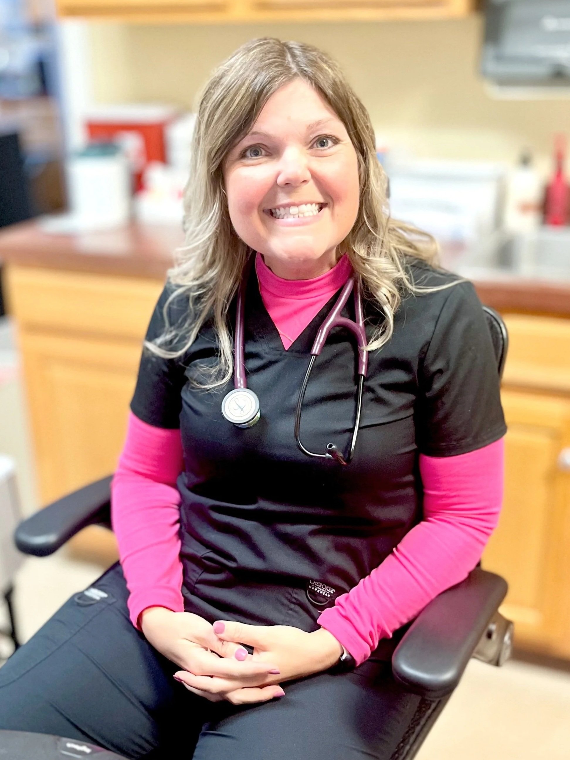 A smiling nurse or medical professional sitting in a chair, wearing black scrubs with a pink long sleeve shirt underneath, and a stethoscope around her neck.