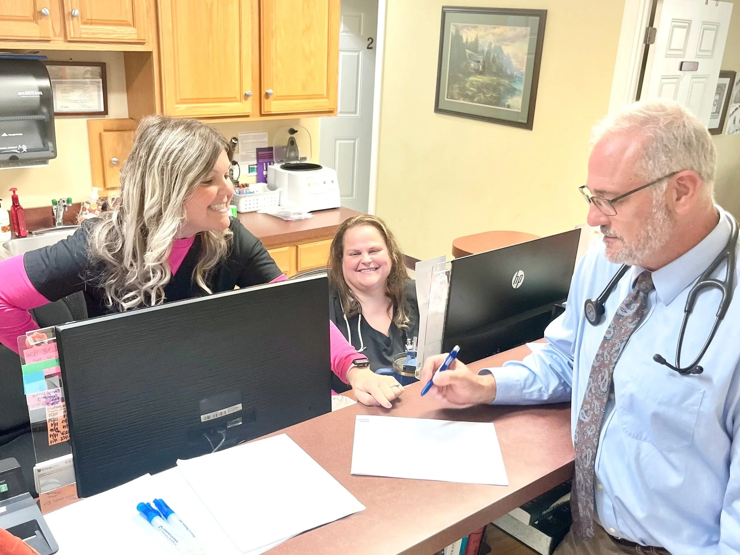 A doctor with glasses and a stethoscope around his neck, sitting at a desk while writing on a sheet of paper, talking to a woman in black clothing, with another woman sitting between them smiling.