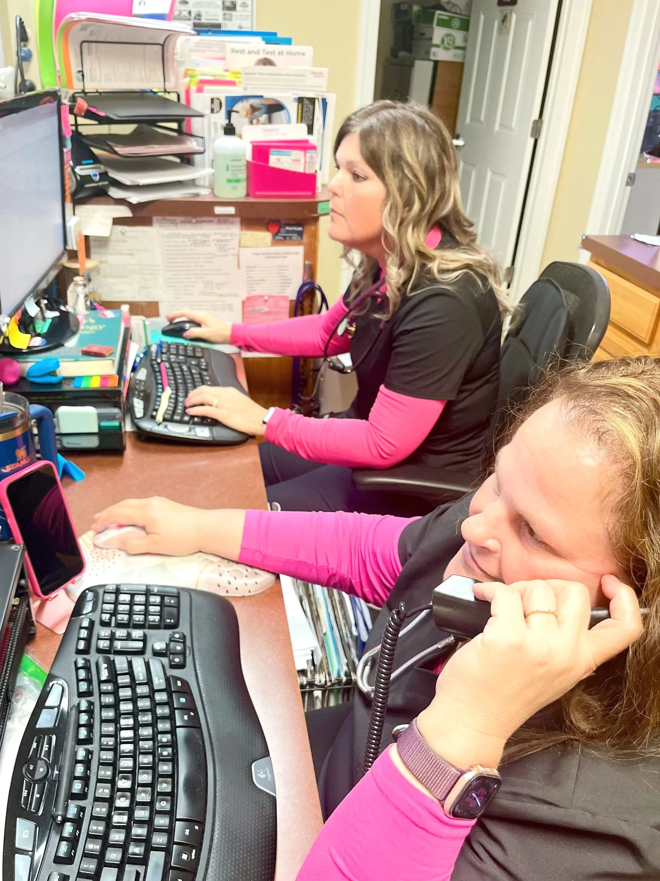 Two women, dressed in black scrubs with pink sleeves, working at a desk in a medical office, using computers and phones. The workspace has various office supplies, a computer monitor, and wall notes.
