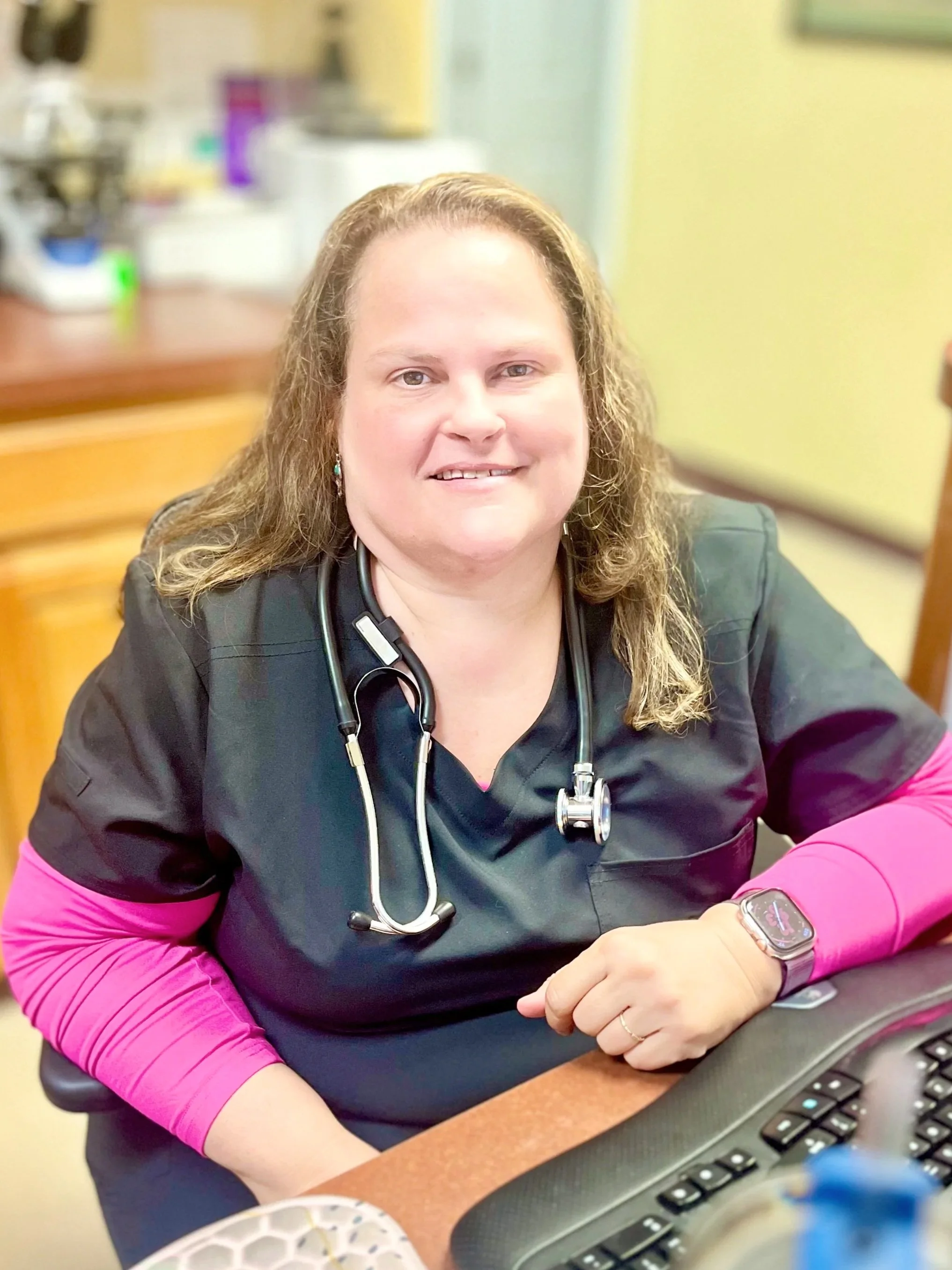 A woman in medical scrubs with a stethoscope around her neck, sitting at a desk in a medical office.