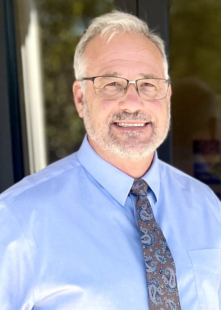 A smiling middle-aged man with gray hair, glasses, a beard, wearing a light blue dress shirt and a paisley tie, standing outdoors near a glass door or window.