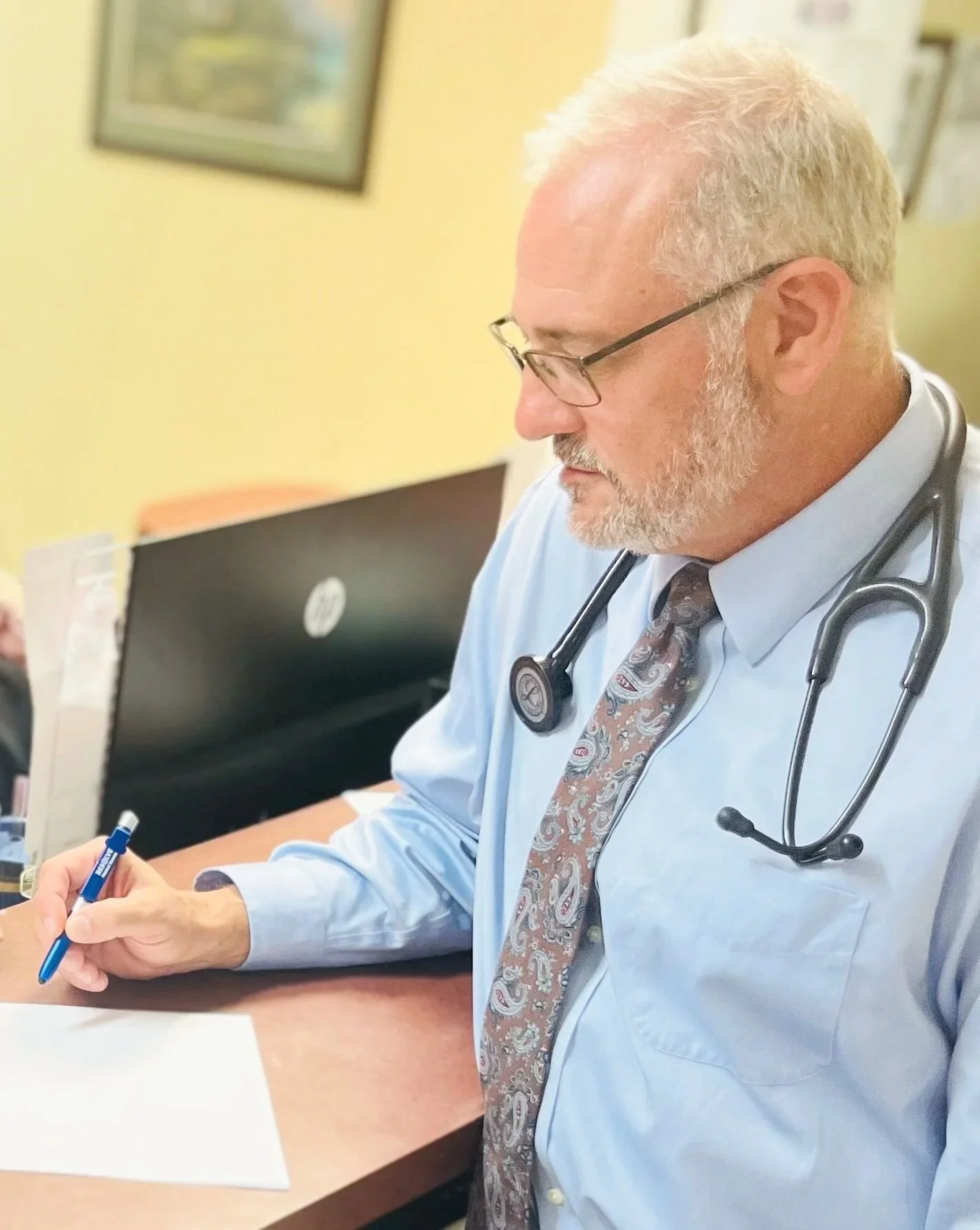A middle-aged male doctor with glasses and a beard, wearing a light blue shirt and a patterned tie, sitting at a desk with a stethoscope around his neck, writing on paper.