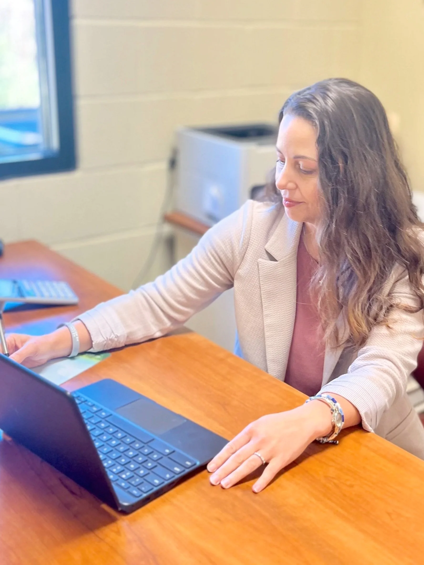 A woman with long, wavy brown hair sits at a wooden desk, working on a laptop with a detached keyboard, in an office setting with a window and a small gray printer in the background.