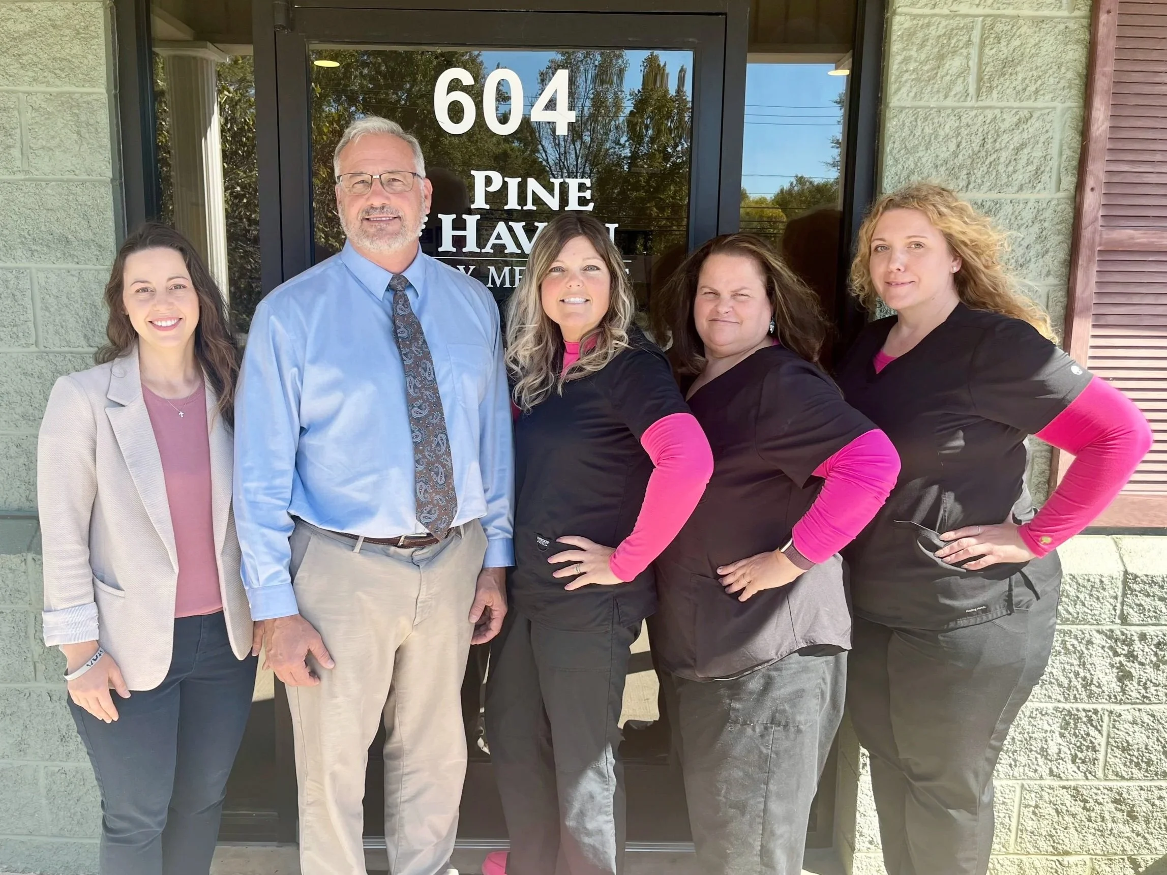 Five people standing in front of a glass door at 604 Pine Haven, smiling outdoors on sunny day. The group includes three women and one man, with one woman on the far left in a beige blazer, men in the center and women on the right wearing black scrubs with pink long sleeves. The background shows trees and a cloudy sky.