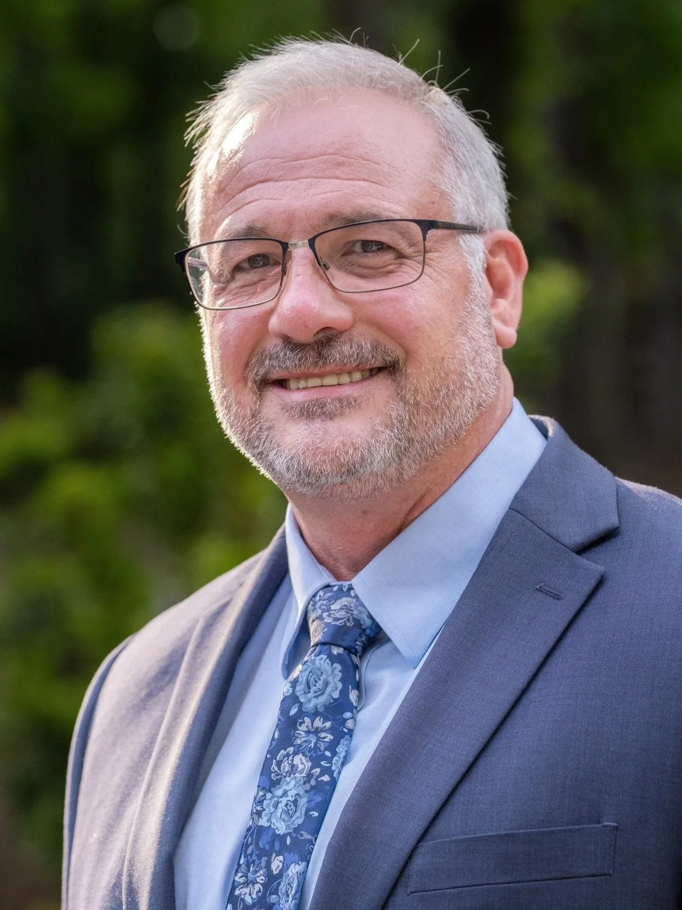 A middle-aged man with glasses, a beard, and short gray hair, smiling outdoors, wearing a blue jacket, light blue shirt, and a floral tie, with a wooded background.