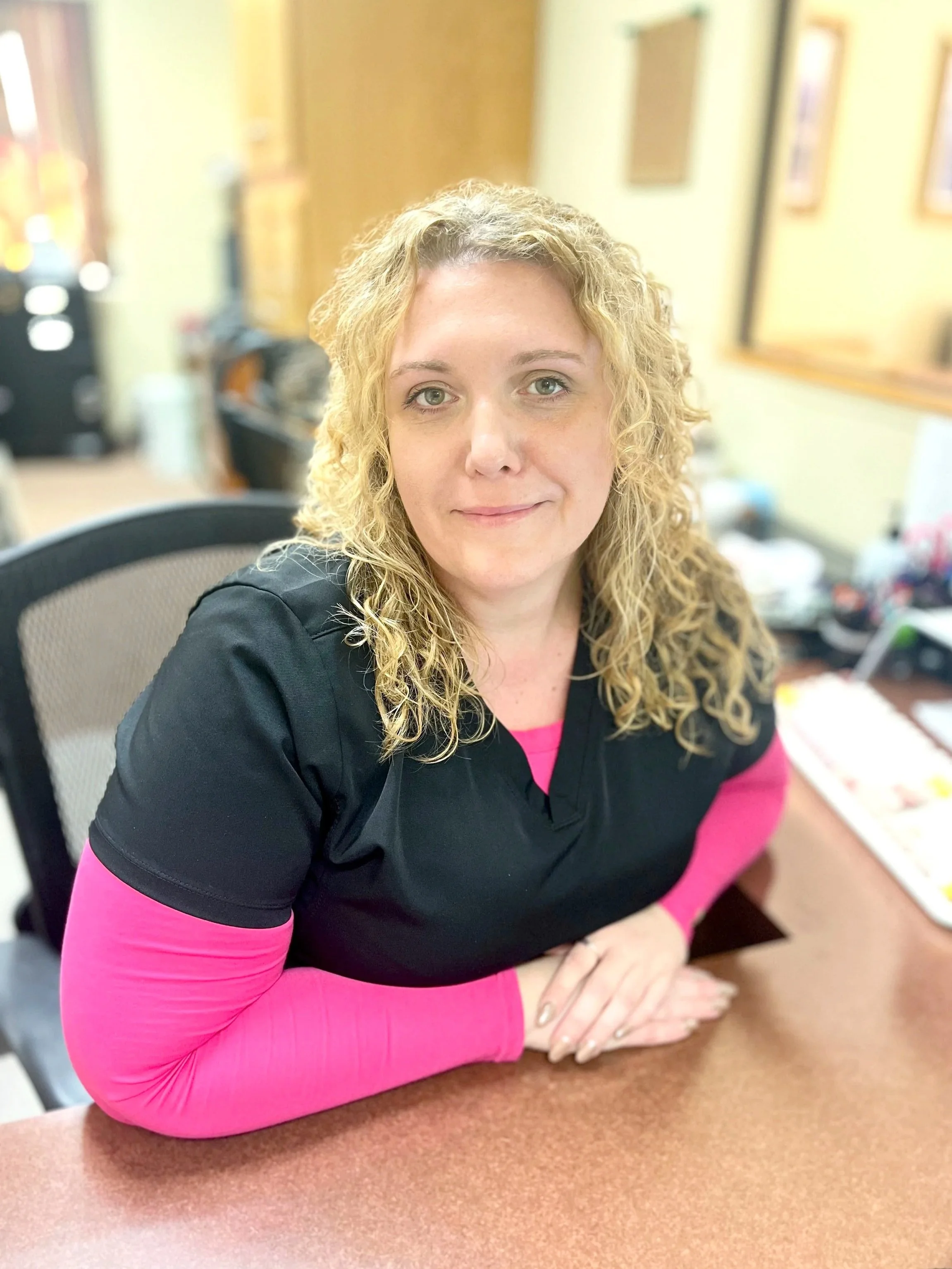 A woman with curly blonde hair sitting at a desk, wearing a black top with pink long sleeves underneath, in an office setting.
