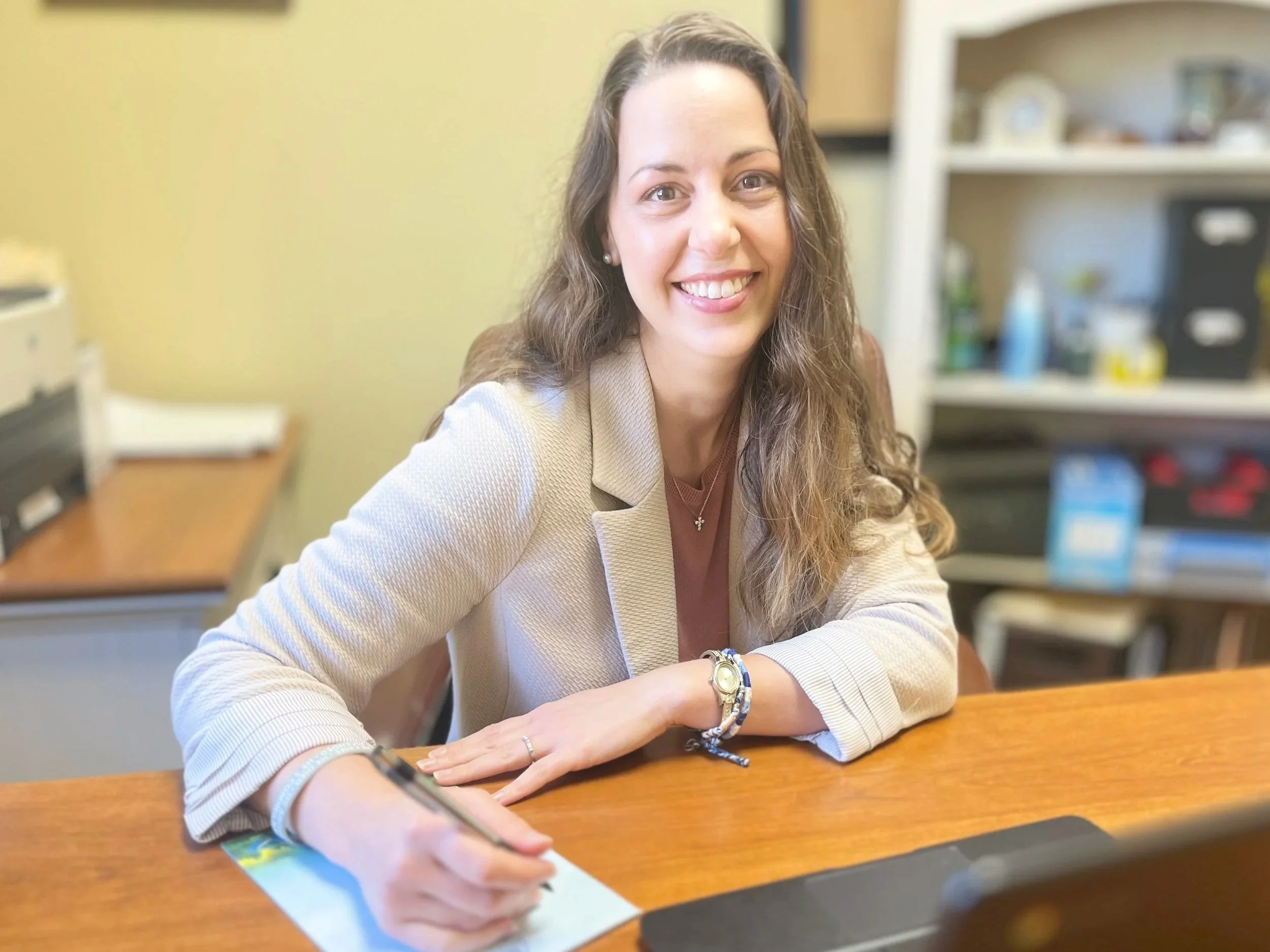 A woman with long brown curly hair smiling and sitting at a wooden desk with a pen and notepad, in a room with shelves and office supplies in the background.