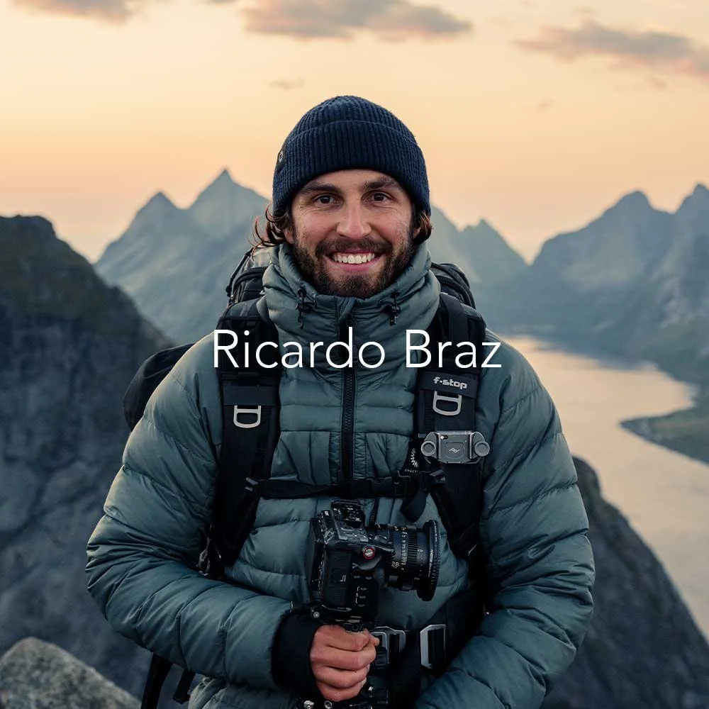 Ricardo wearing a blue beanie and gray jacket hiking with a camera around his neck, standing outdoors in a mountainous landscape at sunset.