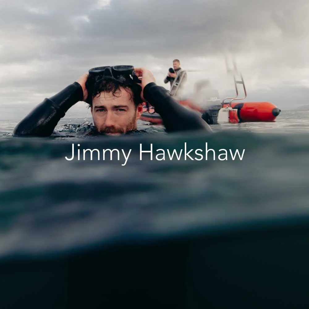 A man in a wetsuit holding a diving mask, partially submerged in water, with a boat and another person in the background on a cloudy day. The text says 'Jimmy Hawkshaw.'