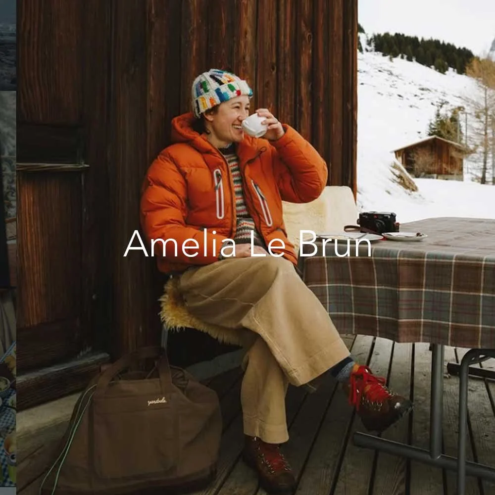 Amelia in an orange jacket and colorful knit hat sits outdoors, smiling and drinking from a mug, with snowy mountains and a wooden cabin in the background.