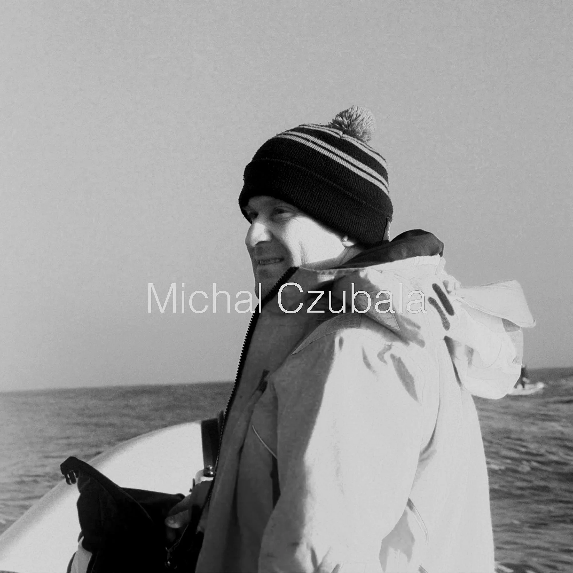 A man in a winter jacket and a striped beanie hat standing on a boat at sea, holding a surfboard.