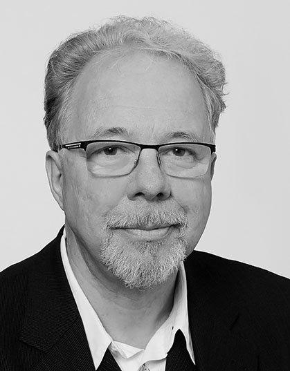 Porträt eines älteren Mannes mit Brille, grauem Haar und Bart, in formeller Kleidung, vor hellem Hintergrund.