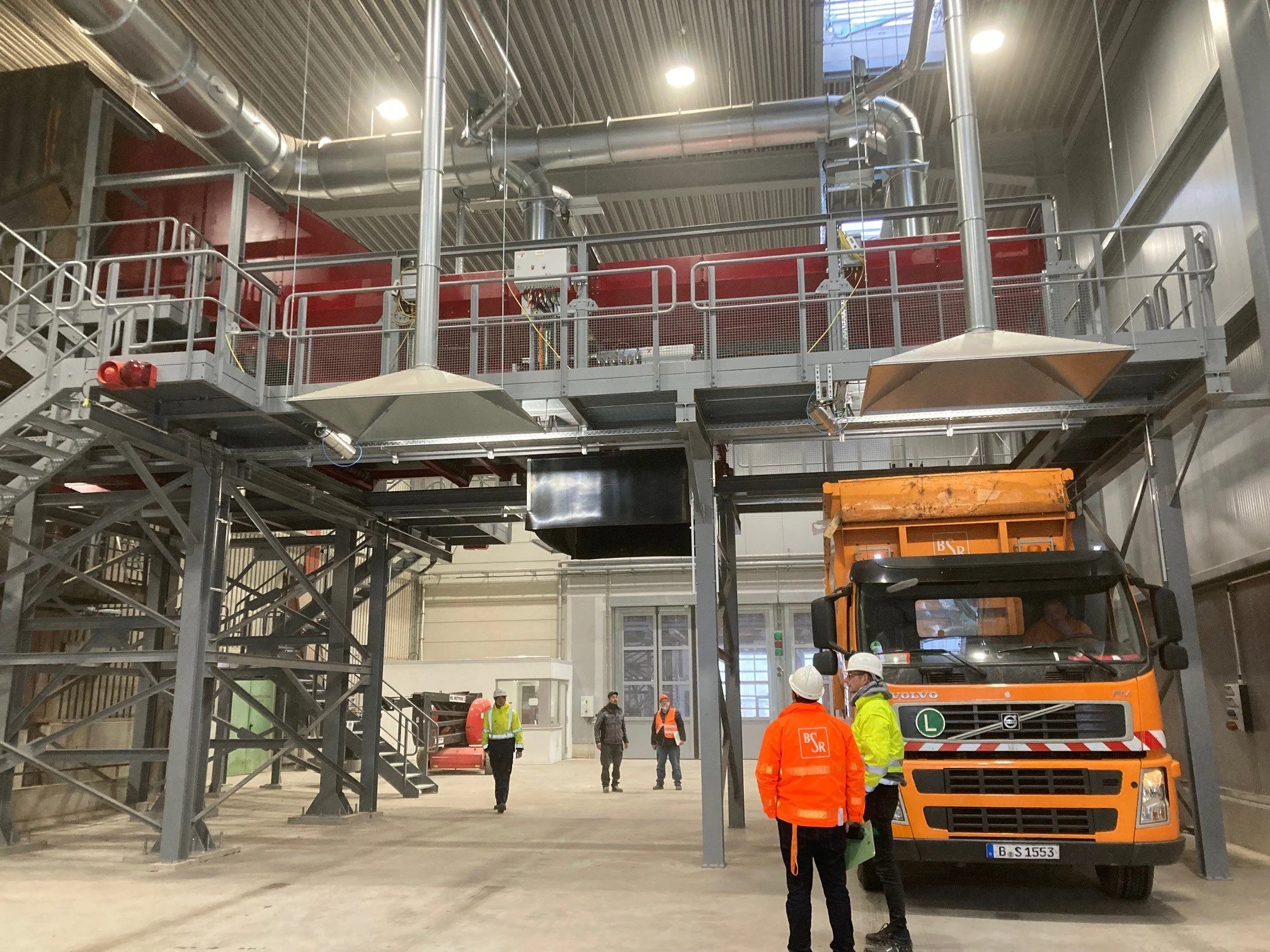Innenansicht einer Industriehalle mit erhöhtem Arbeitsbereich, mehreren Arbeitern in Schutzkleidung und einem orangefarbenen LKW.