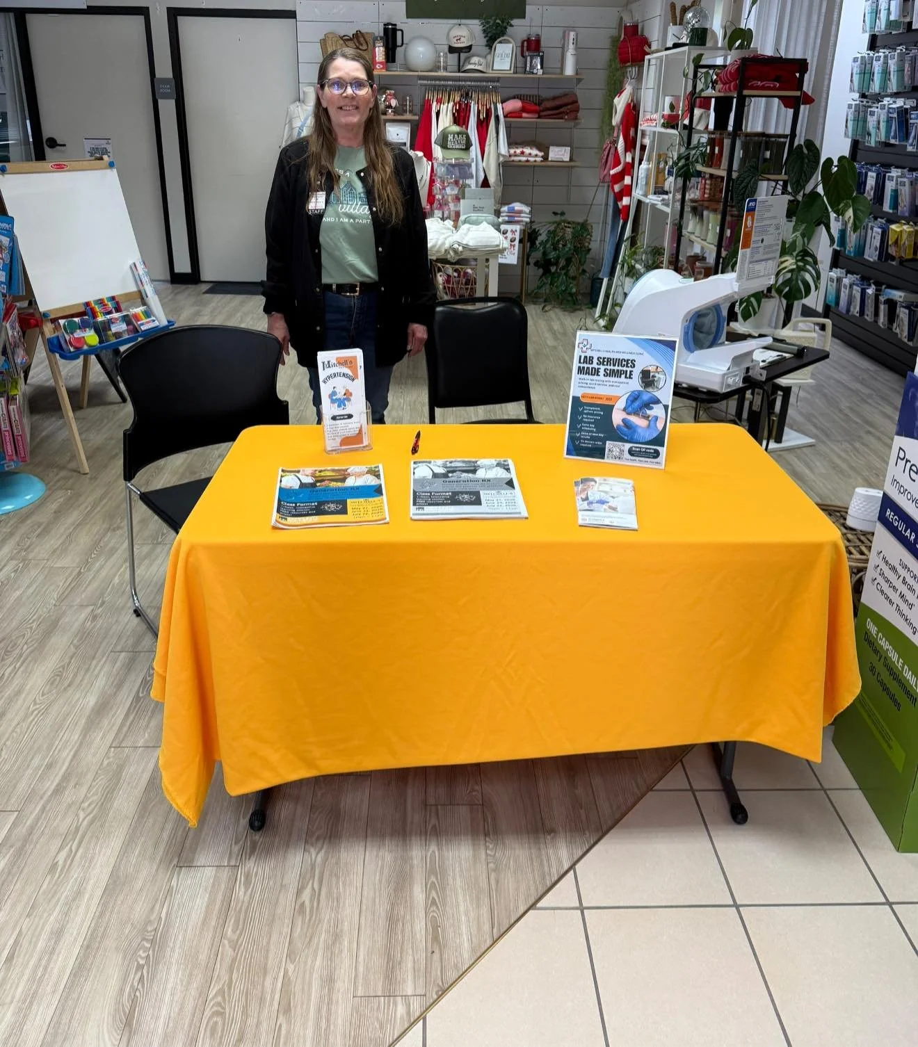 A woman stands behind a table draped with a yellow tablecloth, in what appears to be a retail or health clinic setting. The table has informational pamphlets and a sign about lab services. Shelves with products and clothing are visible in the backgro
