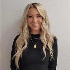 A woman with long blonde hair wearing a black top and gold necklace, smiling at the camera.