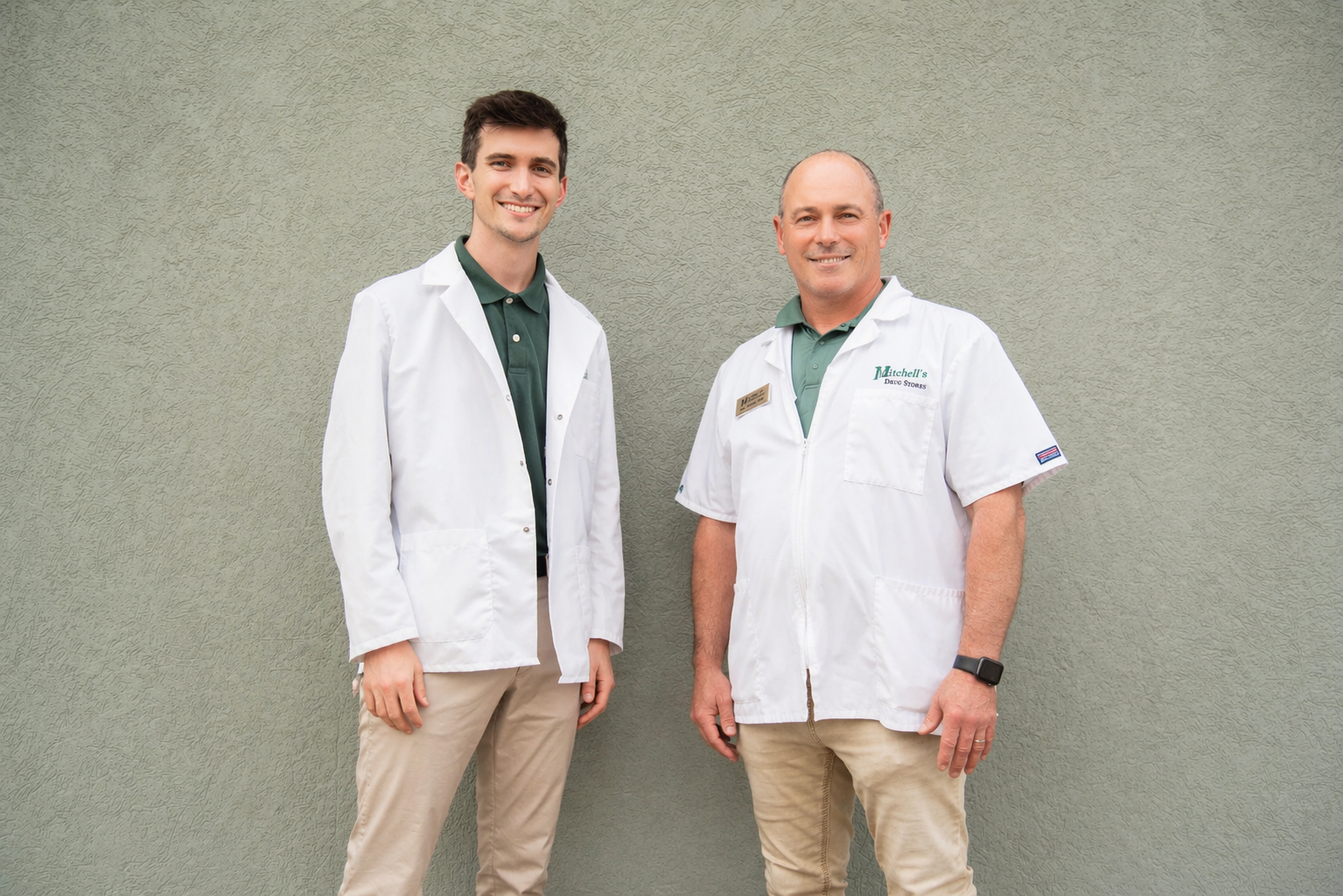 Two smiling men in white medical coats standing against a green wall.