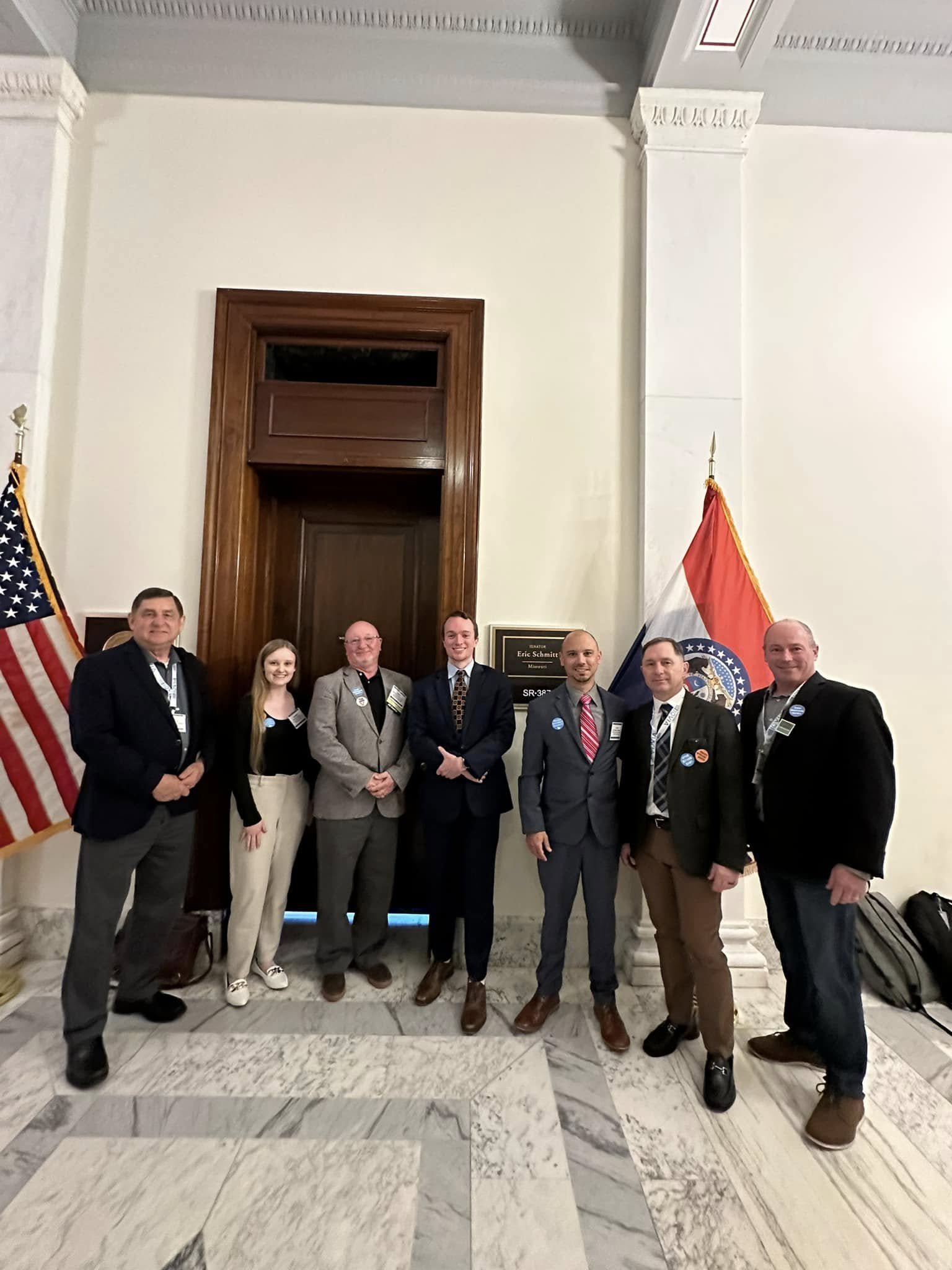 Group of nine people standing in front of a door, with American and Missouri flags on their sides, in a government building with marble floors and white walls.
