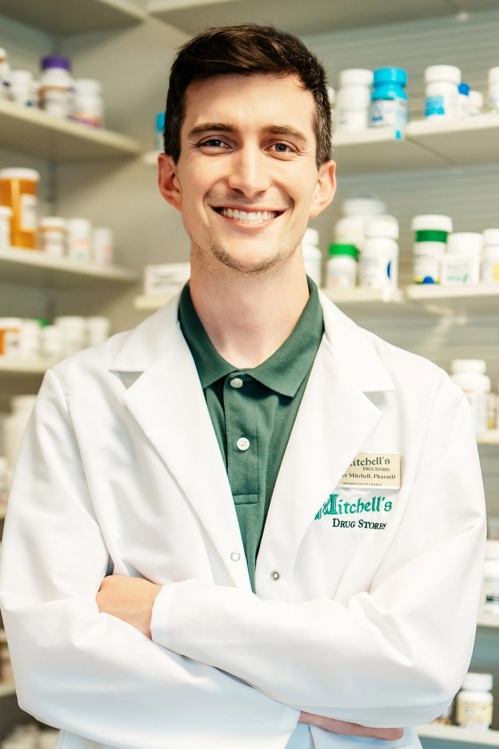 A smiling young male pharmacist in a white coat standing in a pharmacy and crossing his arms.