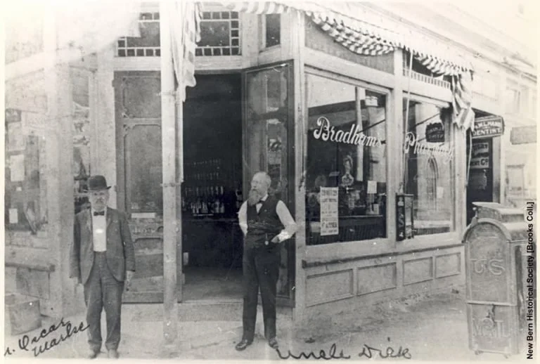 A black and white photograph of two men standing outside a store called Bradham's. The store has large windows displaying bottles and signs. One man is dressed in a suit and bowler hat, while the other wears a vest and shirt. There is an old-fashioned mailbox or postbox to the right of the store entrance, and handwritten notes are visible on the photo.