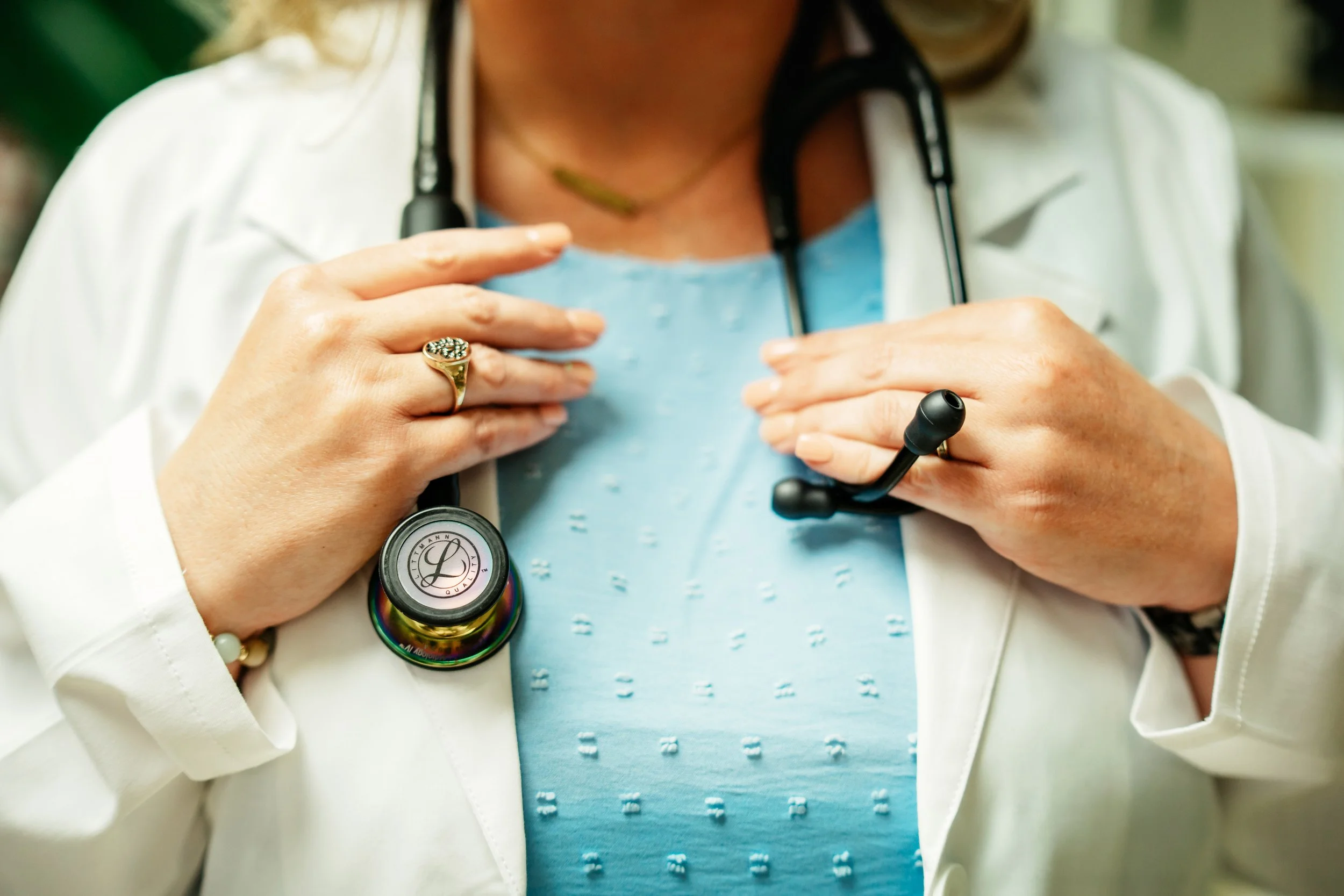 Close-up of a woman in a medical setting wearing a white lab coat and stethoscope, holding her chest with one hand, and wearing a ring on her finger.