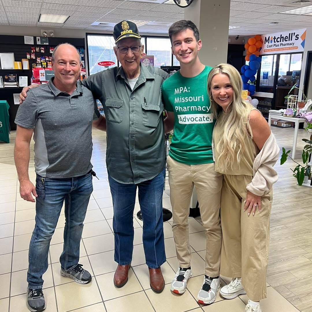 Four people standing inside a pharmacy, smiling at the camera. The group includes two men and two women, with the man in the middle wearing a cap that says 'DRIVE' and an MPA: Missouri Pharmacy Advocate' T-shirt. The background shows shelves of produ