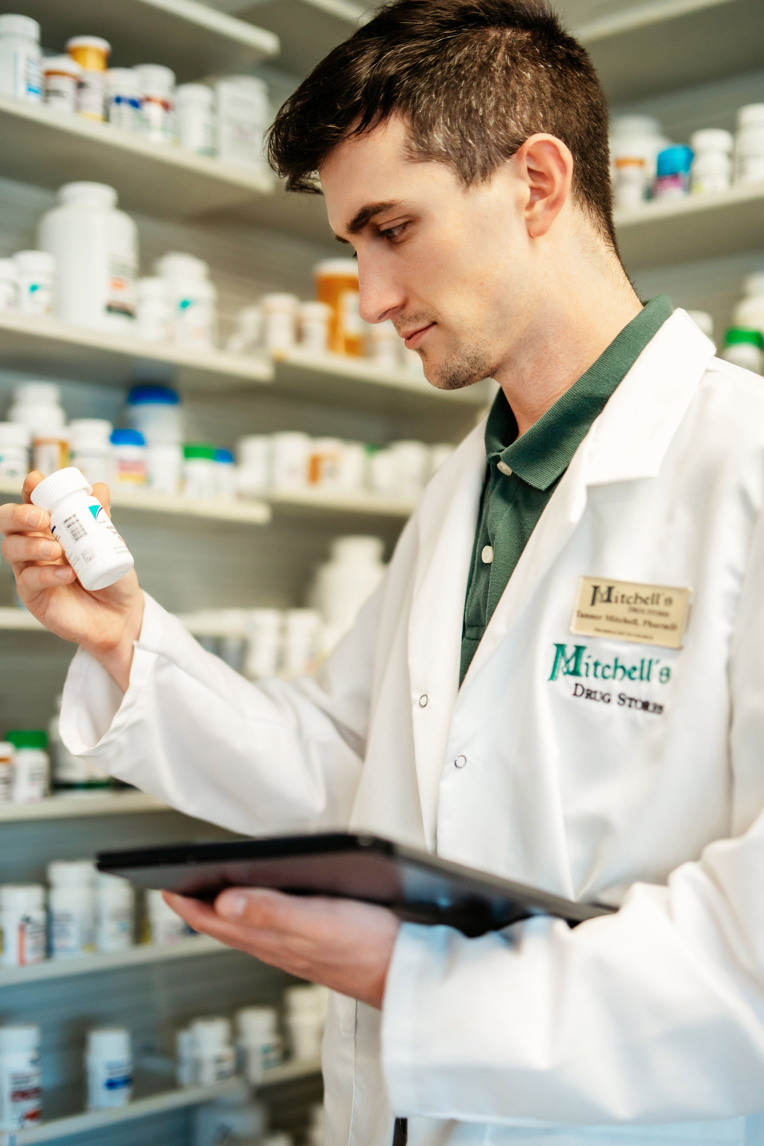 A young man wearing a white coat with a name tag that says 'Mitchell's Drug Store' is looking at a pill bottle in his right hand while holding a tablet device in his left hand, with shelves of various pill bottles in the background.