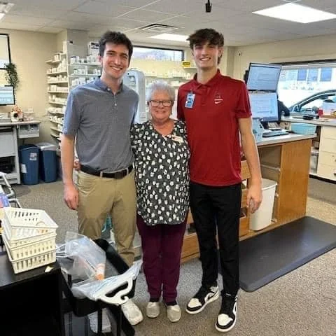 Three people, two young men and an older woman, standing together inside a pharmacy or medical office. They are smiling and posing for the photo.