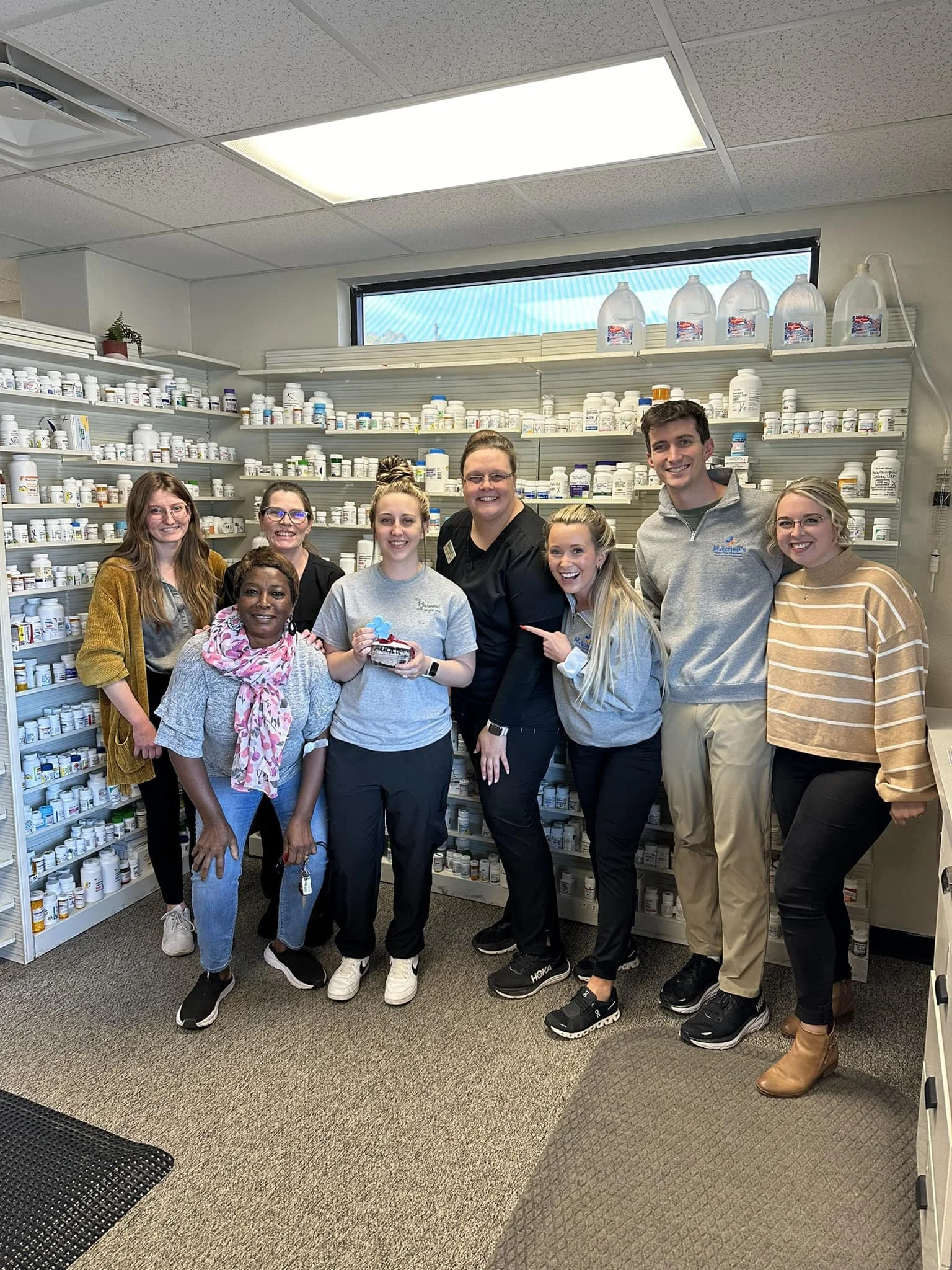 Group of nine people standing in front of pharmacy shelves filled with medication bottles, smiling at the camera.