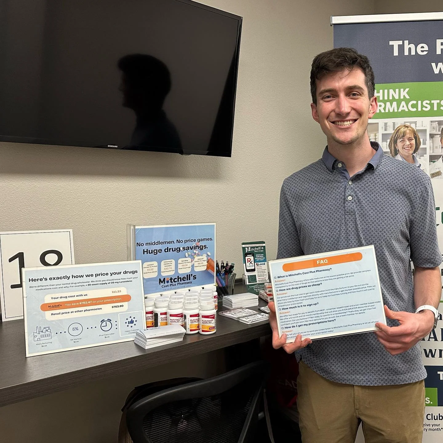 A smiling man holding an informational flyer at a pharmacy booth. The booth displays medication bottles, informational posters, and signage about drug savings and pharmacy services.