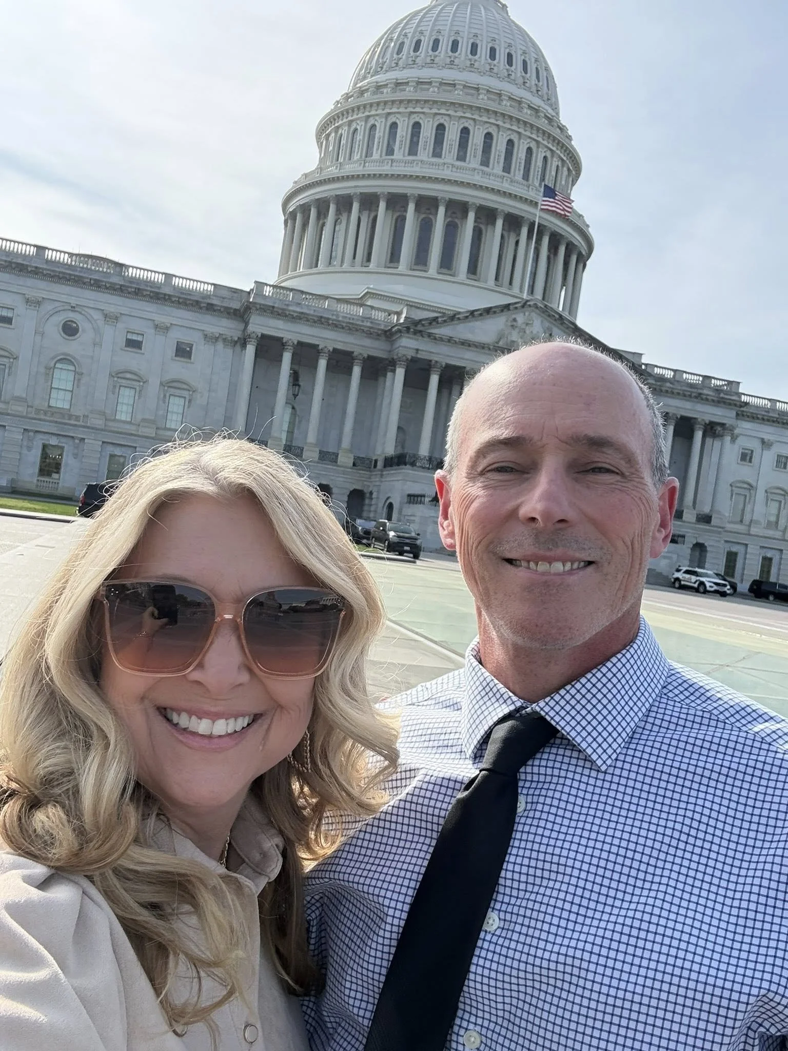 A smiling blonde woman wearing sunglasses and a beige coat taking a selfie with a smiling man in a checkered shirt and black tie in front of the United States Capitol building, with American flag visible on the building.
