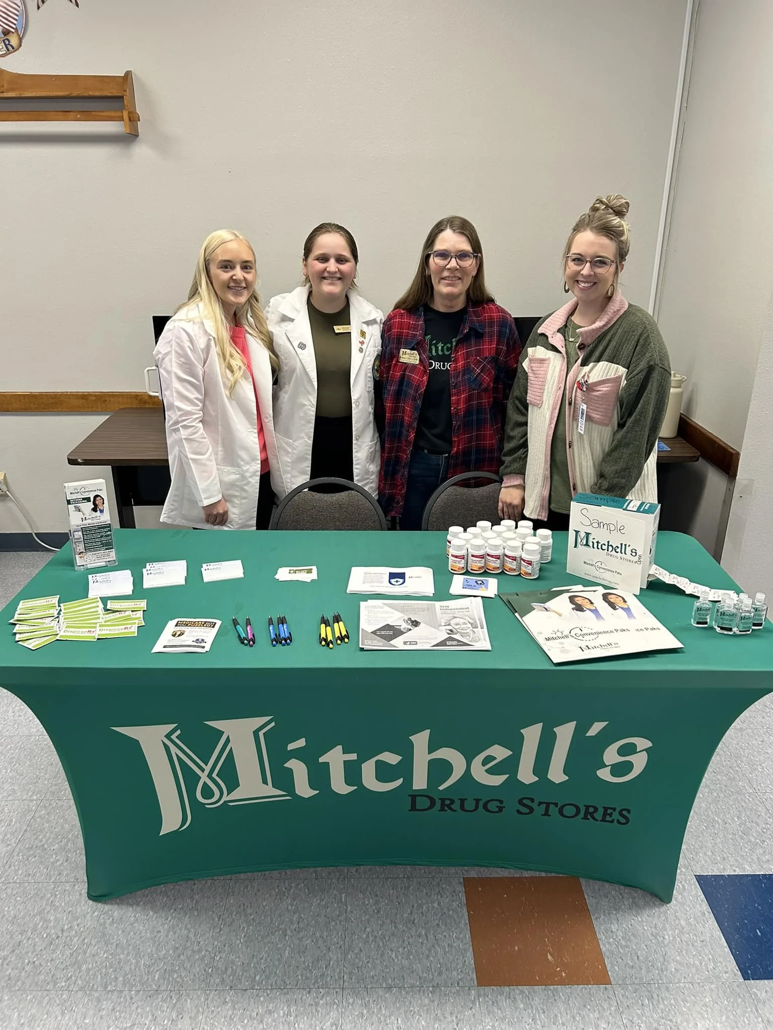 Table with informational pamphlets, bottles, and promotional materials for Mitchell's Drug Stores, with four women posing behind it.