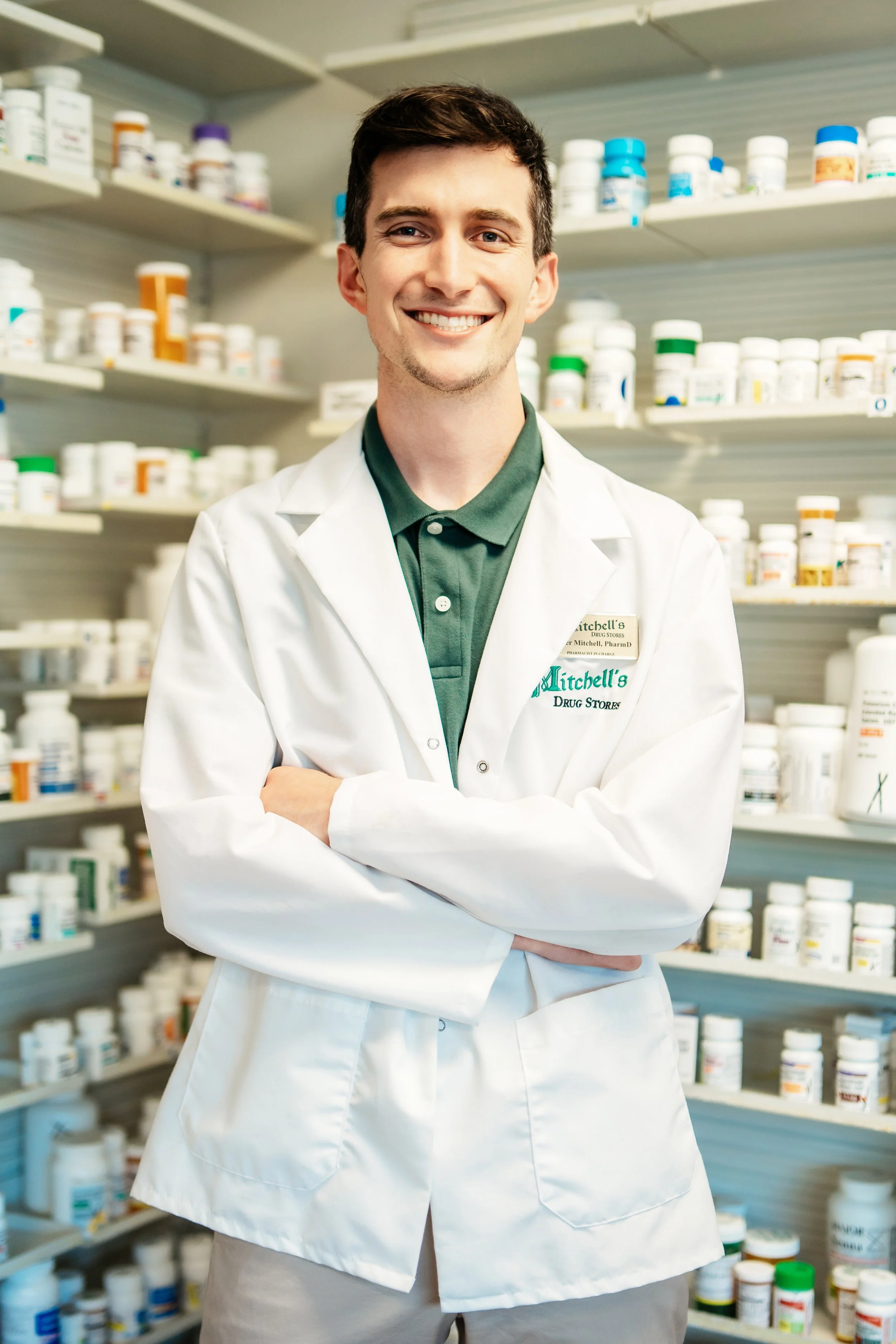 A young male pharmacist wearing a white lab coat standing with arms crossed in front of shelves filled with prescription medicine bottles.